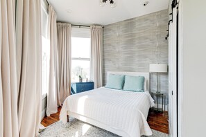 A bedroom with a bed, curtains, a nightstand, a lamp, and a view of the outdoors. - Newly Renovated, Spacious Suite with Every Luxury (Richmond)