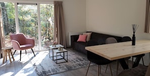 A modern living room with a sofa, a pink chair, a wooden dining table, and a view of a forest through large windows.