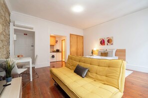 Una sala moderna con un sofá amarillo, una mesa de comedor con sillas, una zona de cocina y una cama con cabecera de madera.