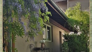 Un jardín con mesa y sillas, rodeado de abundante follaje y flores moradas.