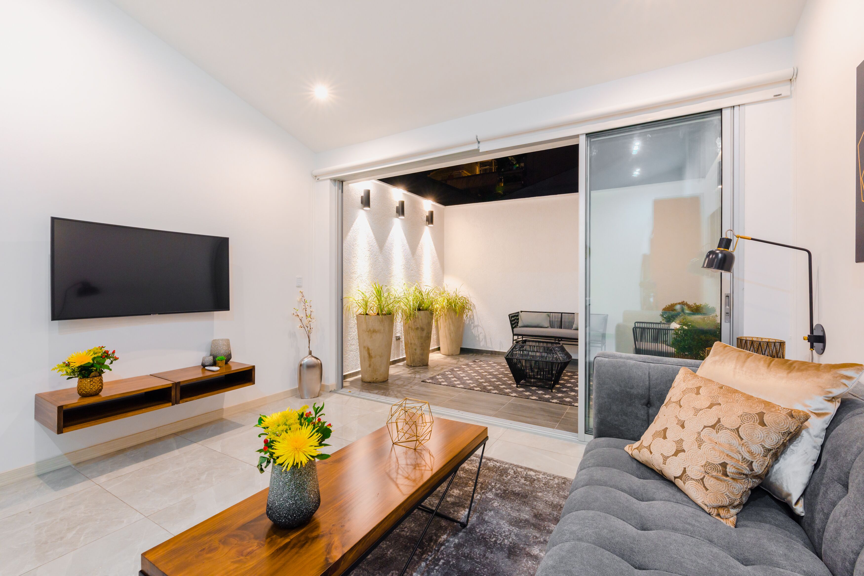 A modern living room with a flat-screen TV, a wooden coffee table with a vase of flowers, a grey sofa with patterned cushions, and a sliding glass door leading to a balcony with potted plants.