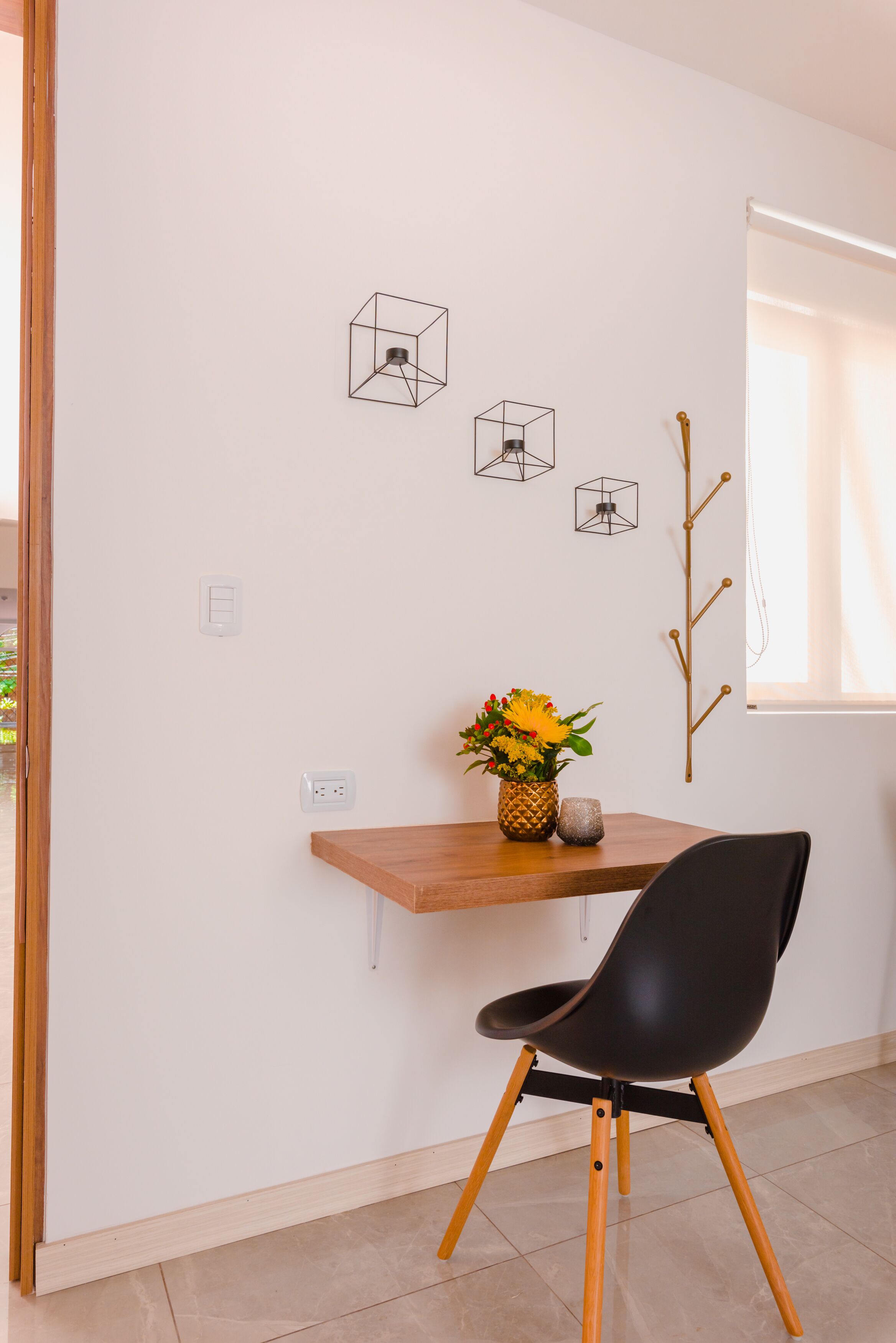 Een moderne kamer met een houten bureau, een zwarte stoel en een vaas met bloemen. Drie geometrische decoraties en een houten tak sieren de wand.