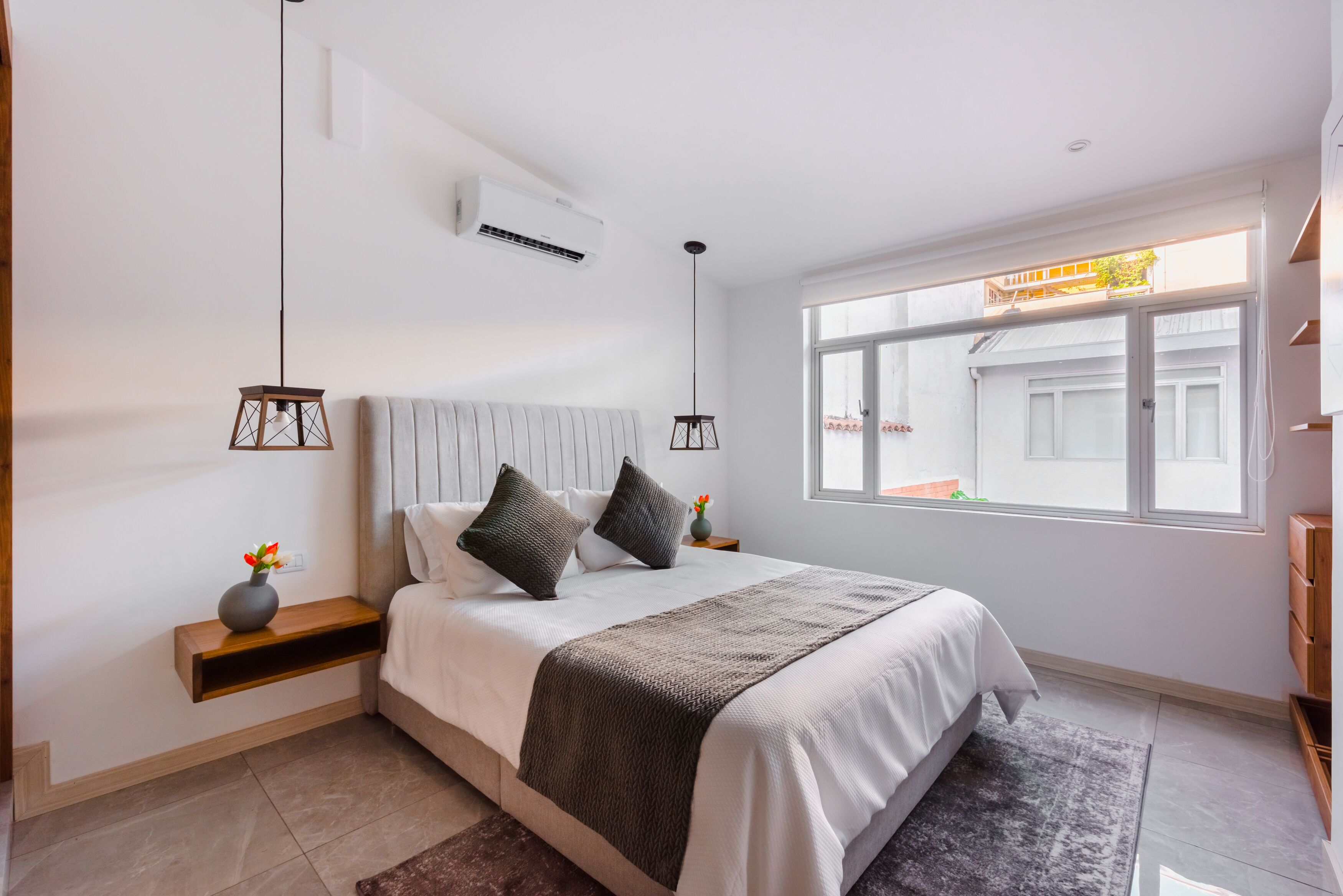 A modern bedroom with a large bed, bedside tables, a hanging light fixture, and a window with a view of the outside.