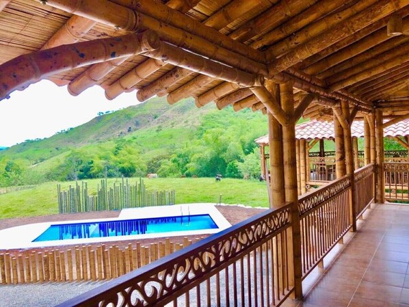 A covered outdoor area with a pool, bamboo pillars, and a view of a green hillside.