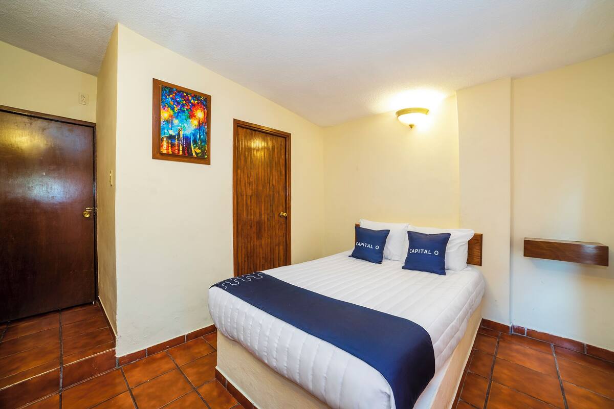 Ein ordentlich bezogenes Bett mit einer weißen und blauen Tagesdecke, zwei Kissen mit dem Aufdruck "Capital O", eine Holztür, ein an der Wand hängendes Bild und eine Wandleuchte.