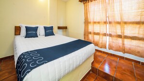 Una cama bien hecha con colcha blanca y azul, dos almohadas a juego y una manta azul. Tiene un cabecero de madera, una lámpara de pared y una ventana con cortina estampada.