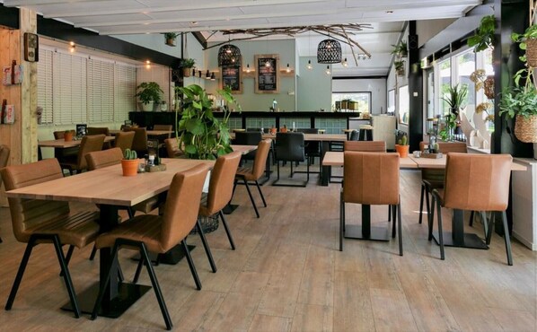 A modern restaurant with wooden tables and chairs, potted plants, and a bar area.