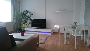 Ein modernes Wohnzimmer mit Flachbildfernseher, Esstisch mit Stühlen, einem Blumentopf und einem kleinen Teppich.