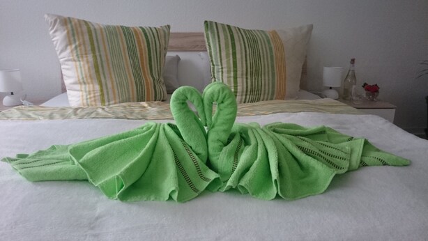 Ein ordentlich bezogenes Bett mit gestreiften Kissen und einem grünen Handtuch, das zu einem Herz geformt ist.