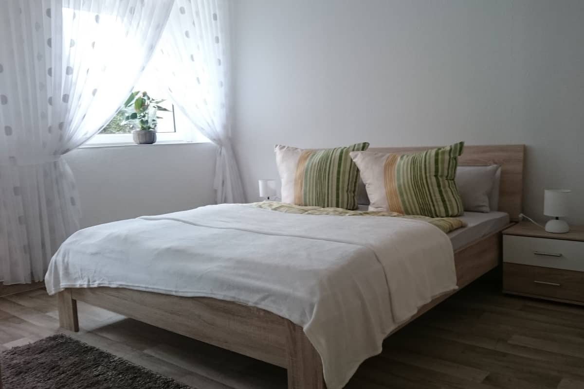 A bedroom with a wooden bed, a bedside table with a lamp, a nightstand, a window with sheer curtains, and a potted plant.
