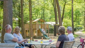 Eine Gruppe von Menschen sitzt an einem Tisch in einem Park, im Hintergrund ist ein Spielplatz zu sehen.