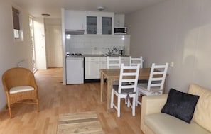A compact living space with a kitchenette, a dining area with white chairs, a wicker chair, and a sofa with a black pillow.