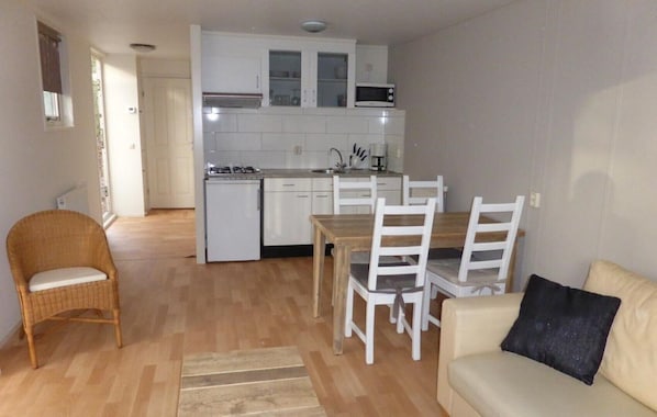 Ein kompakter Wohnraum mit Küchenzeile, einem Essbereich mit weißen Stühlen, einem Korbstuhl und einer Couch mit schwarzem Kissen.