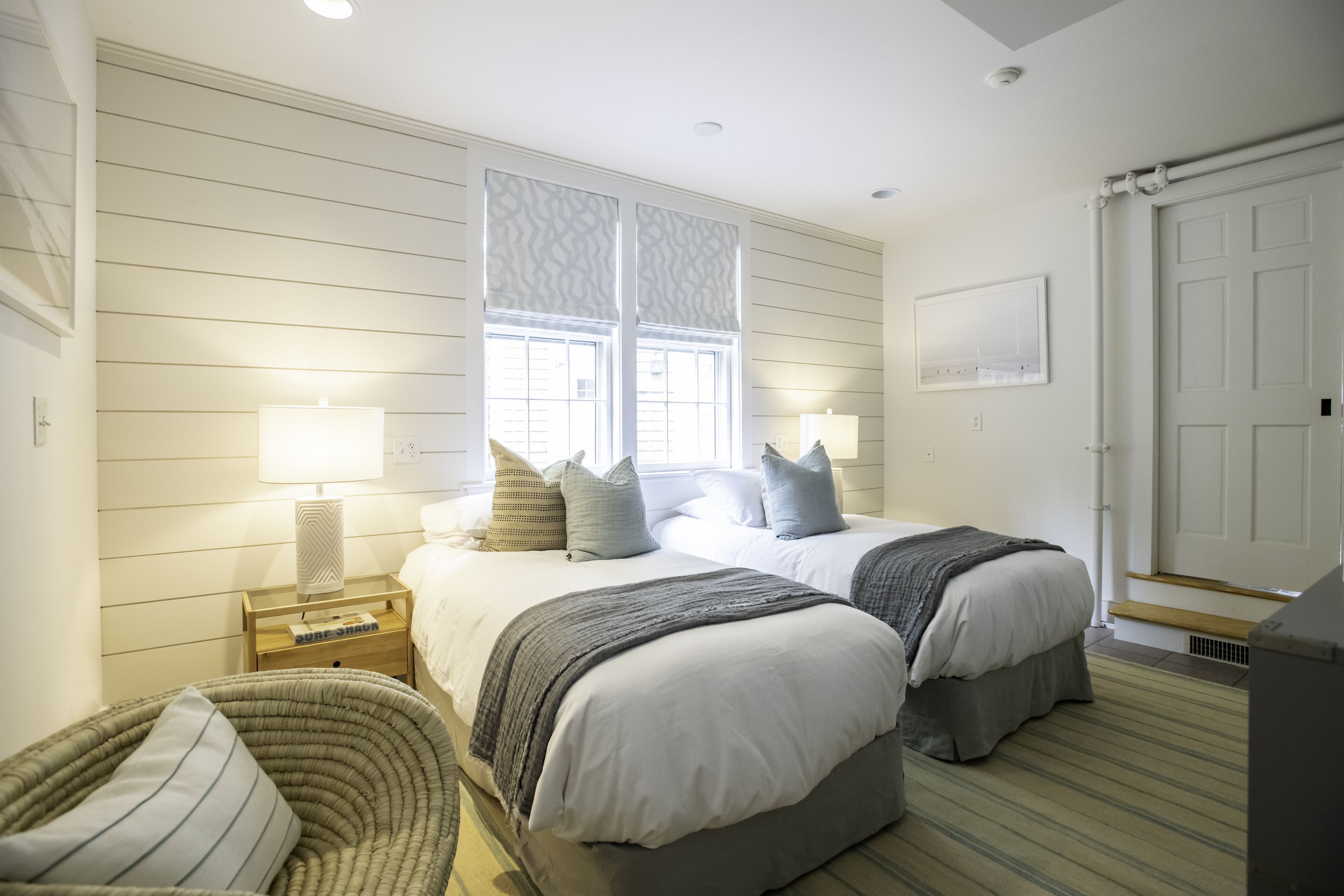 Un dormitorio con dos camas, una ventana con cortinas estampadas, una mesita de noche con lámpara y una silla.