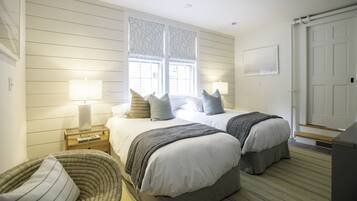 Un dormitorio con dos camas, una ventana con cortinas estampadas, una mesita de noche con lámpara y una silla.