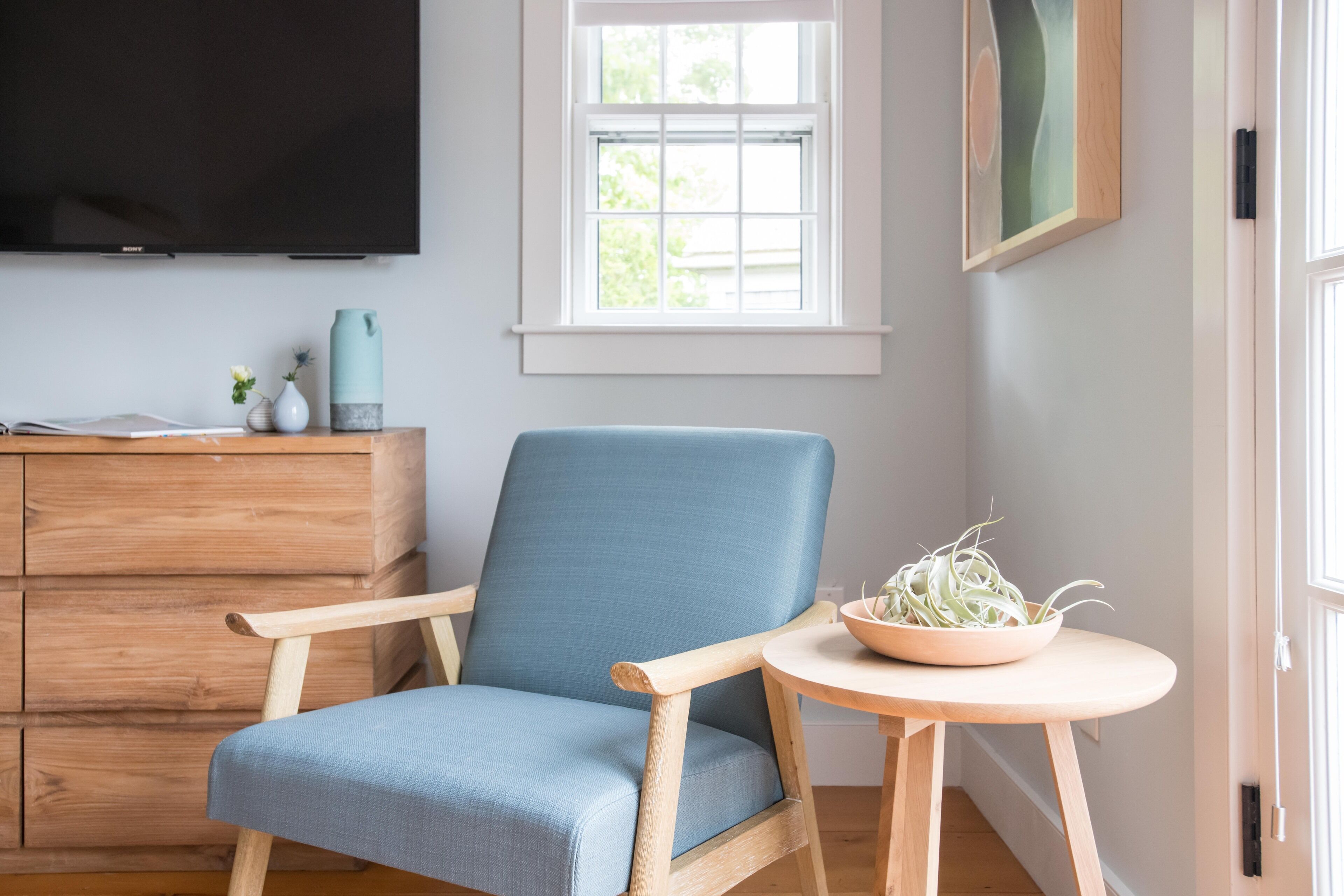 Una habitación con un sillón azul, un aparador de madera, un televisor, una ventana y una mesita con una planta.