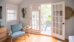 Ein Zimmer mit blauem Sessel, einem kleinen Tisch mit Pflanze, einem Holzschrank und einer Schiebetür zu einem Außenbereich mit Stühlen und einer Terrasse.