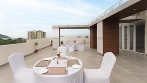 Una terraza en la azotea con mesas redondas dispuestas para comer, sillas blancas y plantas en macetas.