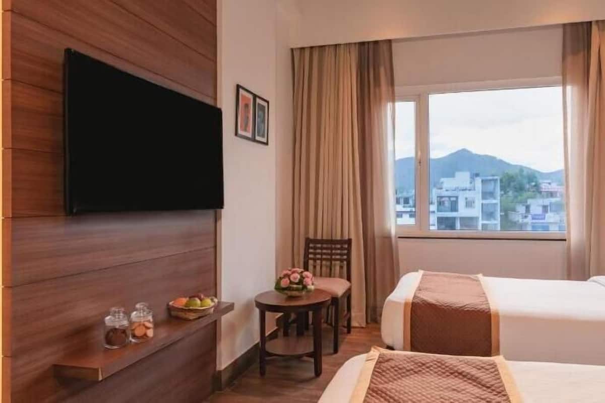 A hotel room with a bed, a TV mounted on the wall, a small table with a vase of flowers, and a view of buildings and mountains through the window.