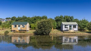 Dos cabañas de madera junto a un lago tranquilo, cada una con una pequeña terraza y una ventana.