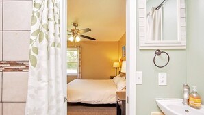 Un dormitorio con cama, ventilador de techo, ventana y baño con lavamanos y artículos de aseo.