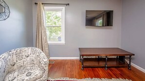 Una habitación con un sillón estampado, una mesa de madera, una ventana con cortinas y un televisor de pantalla plana montado en la pared.