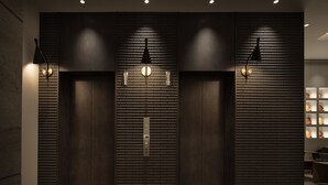 Un vestíbulo moderno con un panel de madera oscura, un ascensor central y dos luces fijadas a la pared.