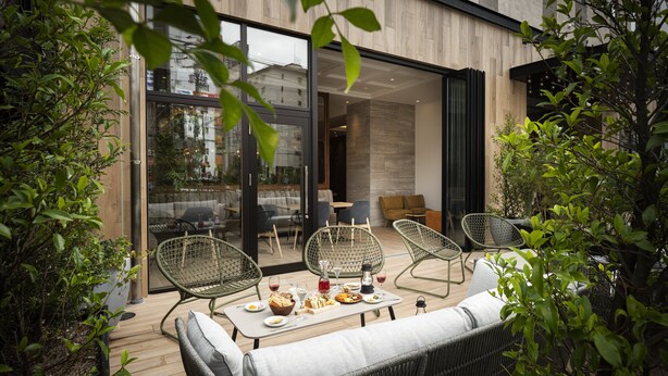 Un área de comedor al aire libre con mobiliario de mimbre, una mesa preparada con comida y bebidas, y vista al interior a través de puertas de vidrio.