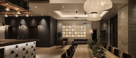 Un espace repas moderne avec une longue table en bois, de nombreuses chaises et des luminaires suspendus. On y trouve un comptoir de cuisine intégré avec un évier et un réfrigérateur. La pièce est agrémentée de lambris en bois foncé, d’un ventilateur de plafond et d’un mur décoré de tableaux encadrés.