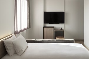 Ein modernes Hotelzimmer mit einem großen Bett, einem flachbildfernseher an der Wand, einer kleinen Holzkommode und einem Korbwäscher.