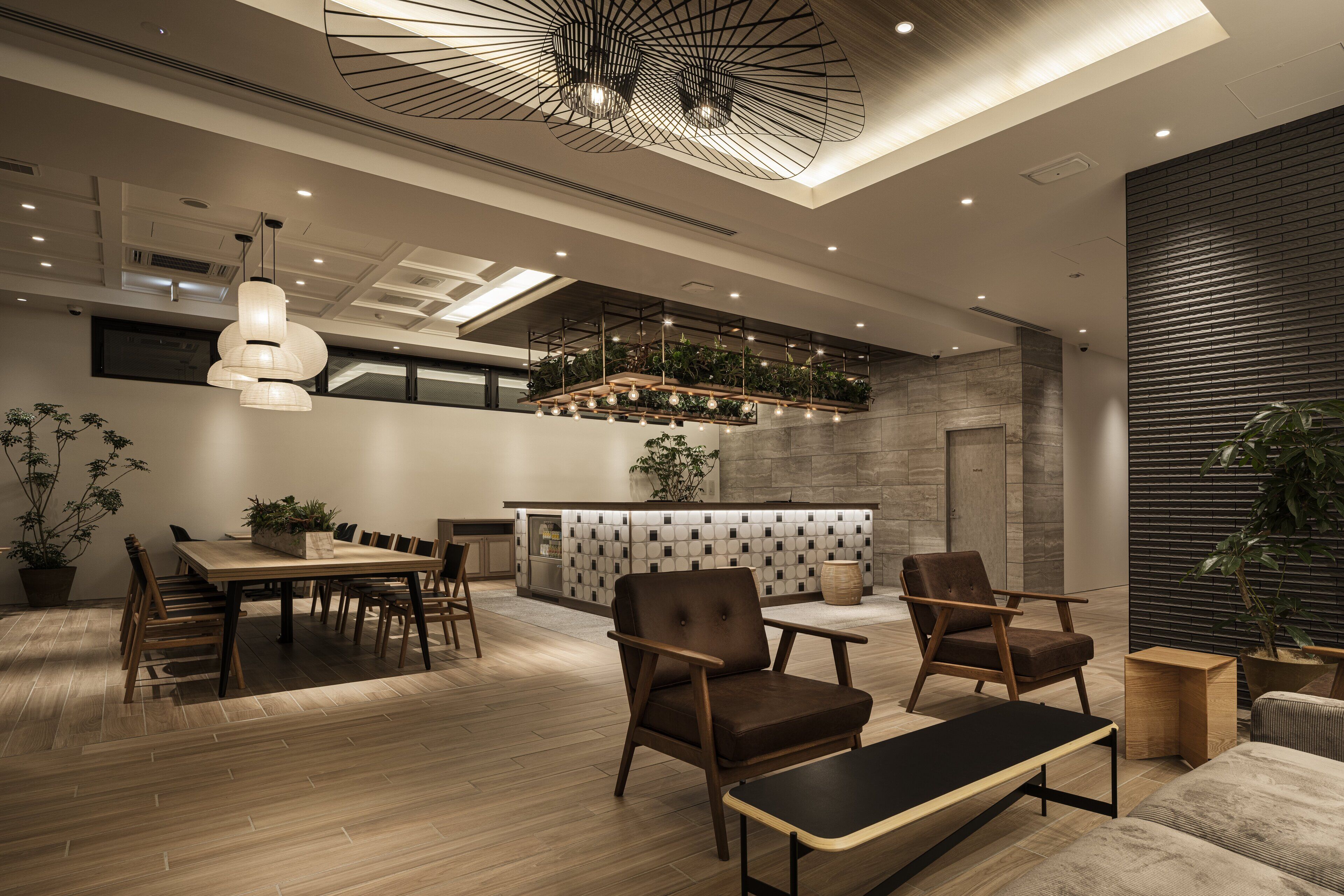 A modern interior with a wooden dining table, chairs, a sofa, and a bench. There is a kitchen area with a countertop and plants.