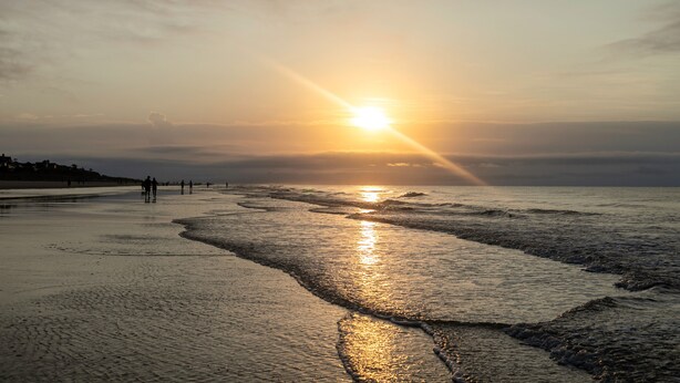 Une plage où le soleil se couche à l’horizon, et où les vagues viennent doucement lécher le rivage.