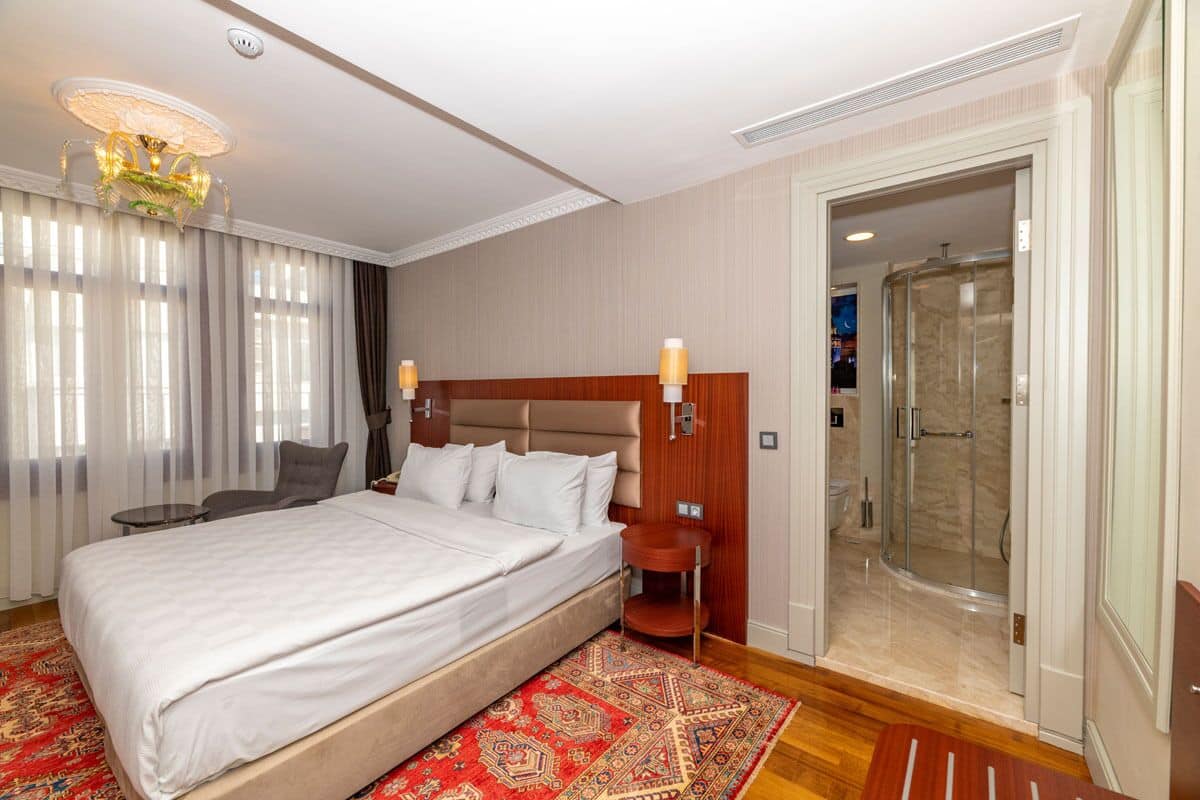 A hotel room with a large bed, a seating area with a chair and table, a shower area, and a chandelier.