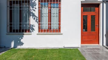 A building with two windows, one with metal bars and one with curtains, and a red door.