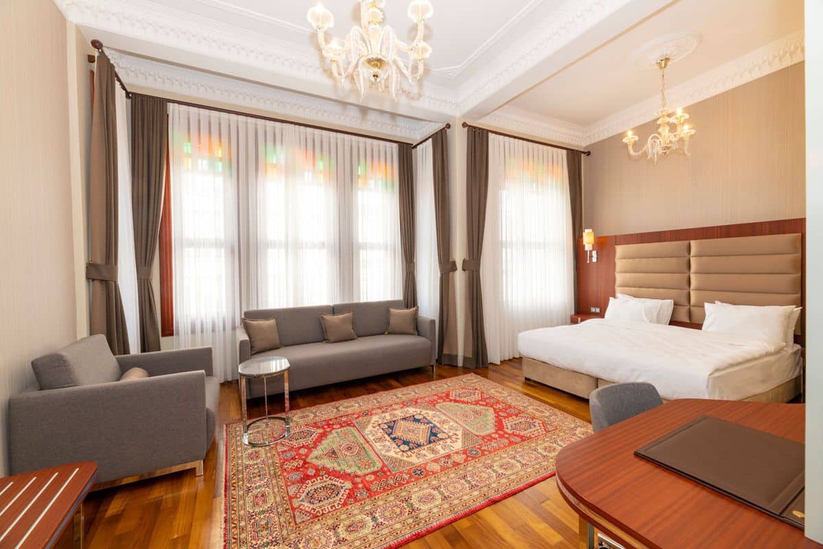 A hotel room with a bed, sofa, armchair, small table, and a large patterned rug.