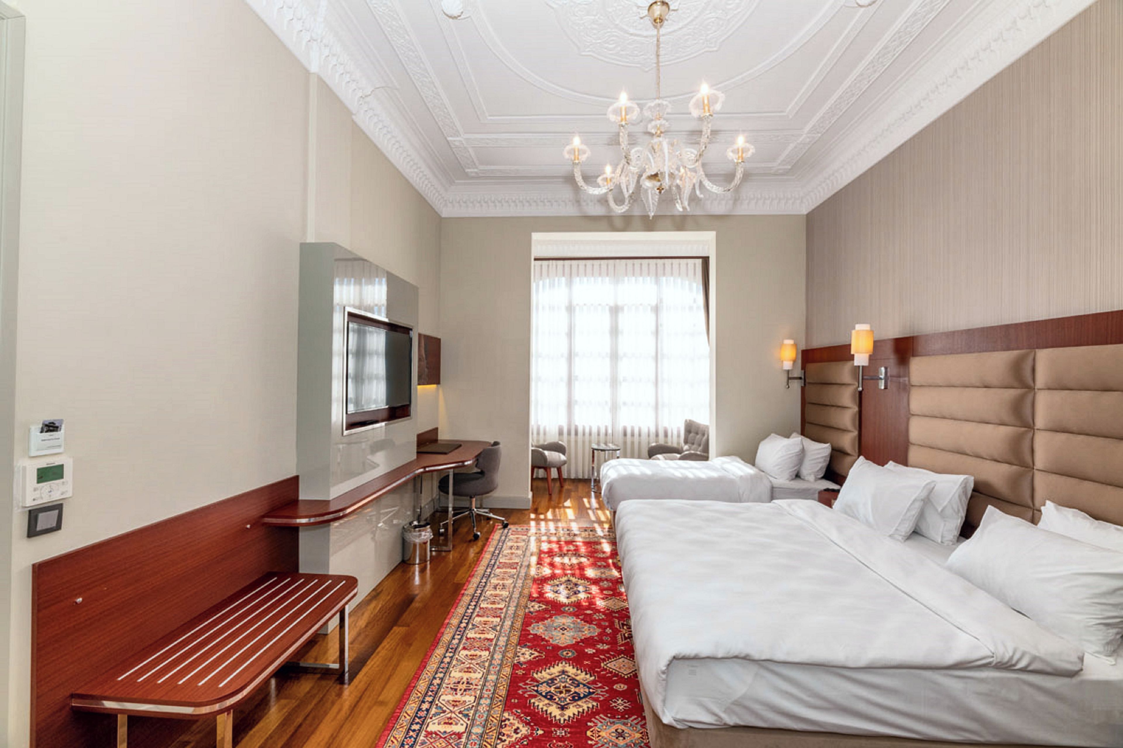 A hotel room with a large bed, a smaller bed, a desk, a television, and a chandelier.