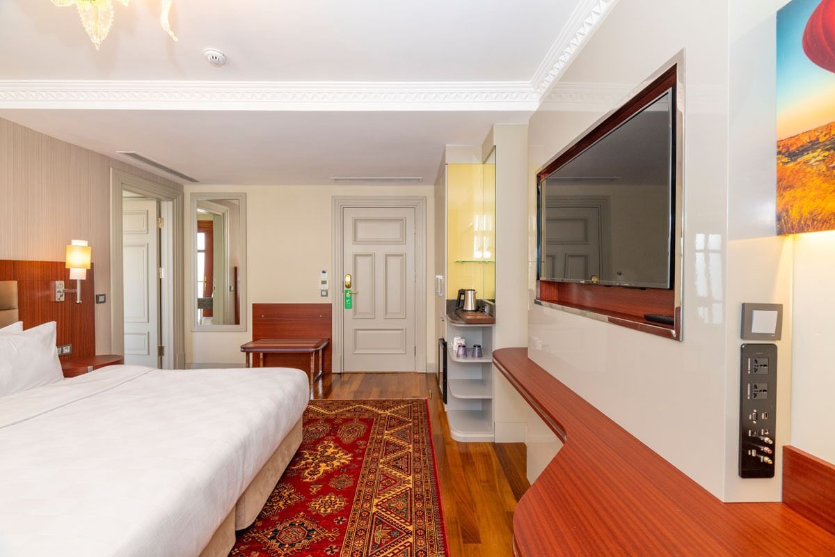Quarto de hotel com cama, escrivaninha, espelho, televisão e painel de controle.