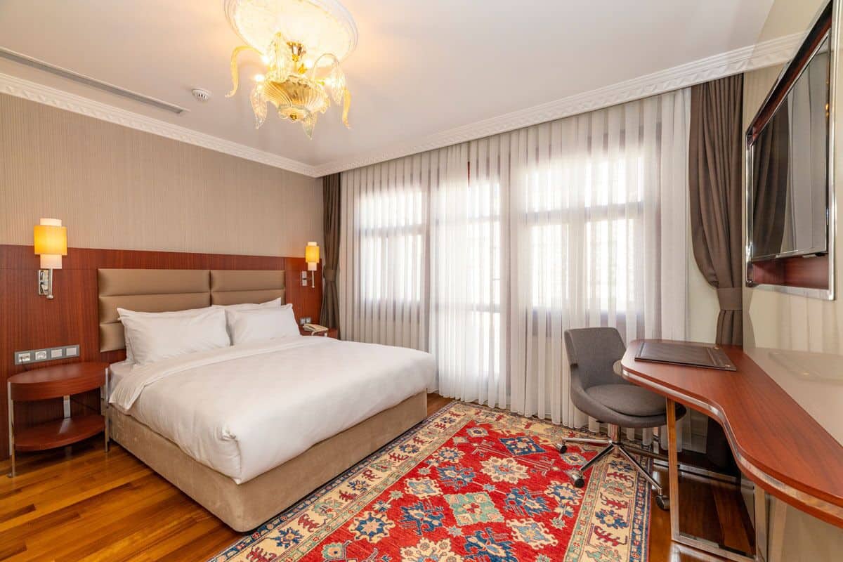 A hotel room with a large bed, a desk, a chair, a chandelier, and a large window with curtains.