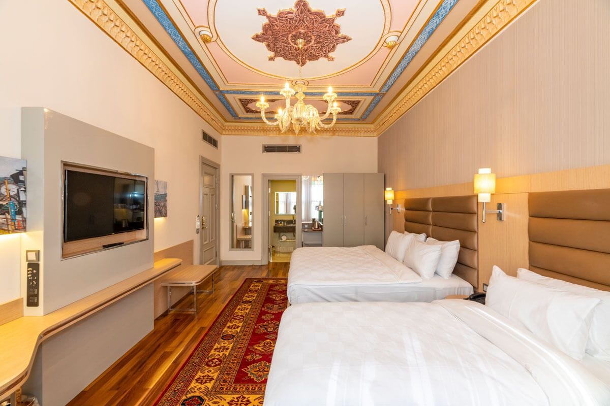 Habitación de hotel con dos camas, televisión, lámpara de araña y alfombra roja.