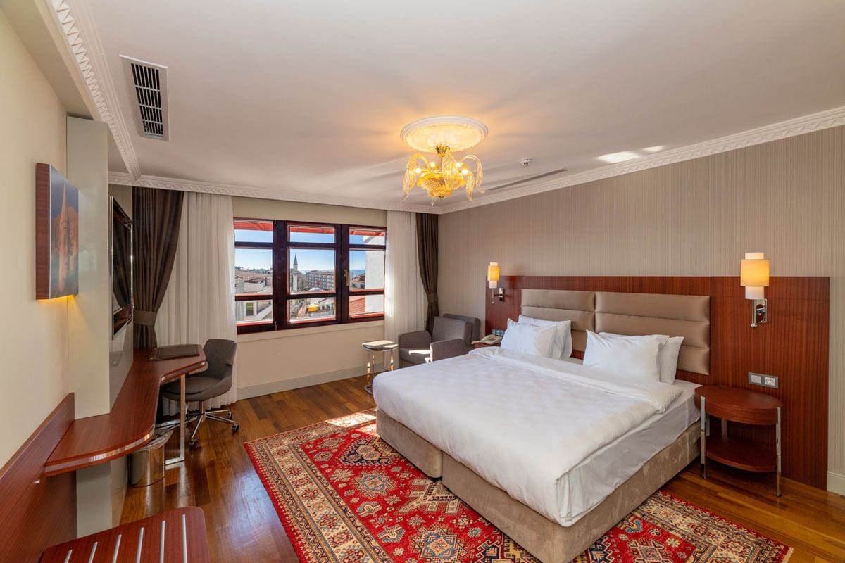 A hotel room with a large bed, a desk, a chair, a nightstand, and a view of the city through the window.