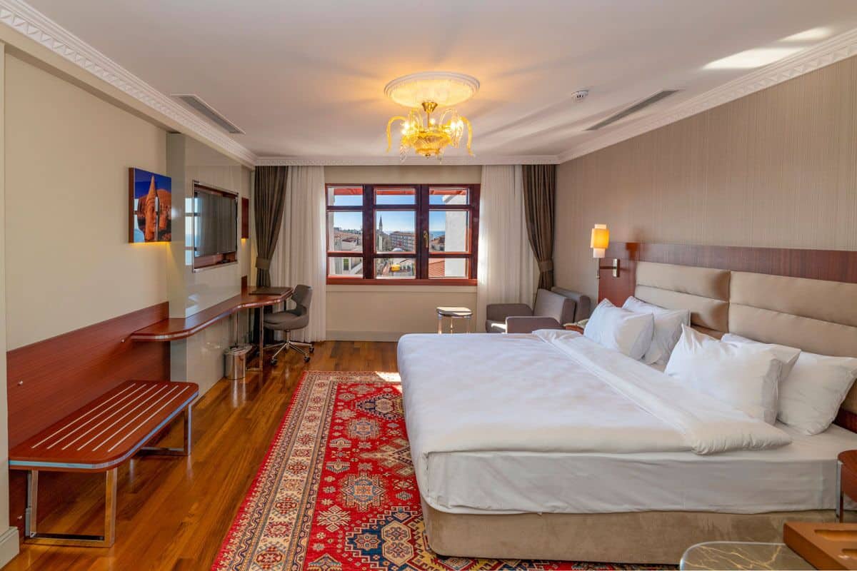 A hotel room with a large bed, a wooden bench, a desk, a chair, a TV, and a window with a view of buildings.