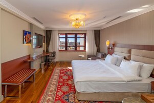 Habitación de hotel con una cama grande, un banco de madera, un escritorio, una silla, un televisor y una ventana con vistas a edificios.