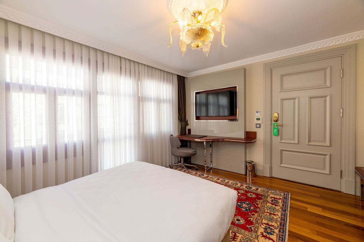 A hotel room with a large bed, a desk with a chair, a television, and a door with a lock.