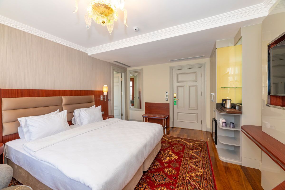 A hotel room with a large bed, a wooden headboard, a red patterned rug, a small table with a lamp, a TV, and a kitchenette area with a microwave and a kettle.