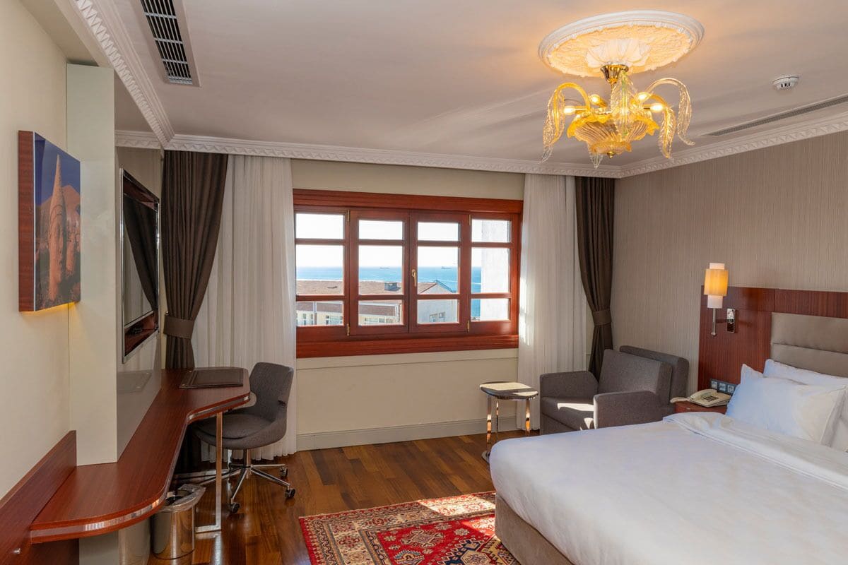 Une chambre d’hôtel avec un lit, un bureau, des chaises, une petite table, un lustre et une fenêtre donnant sur un beau paysage.
