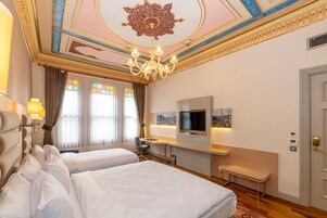 A hotel room with two beds, a desk, a TV, a chandelier, and a large window with curtains.