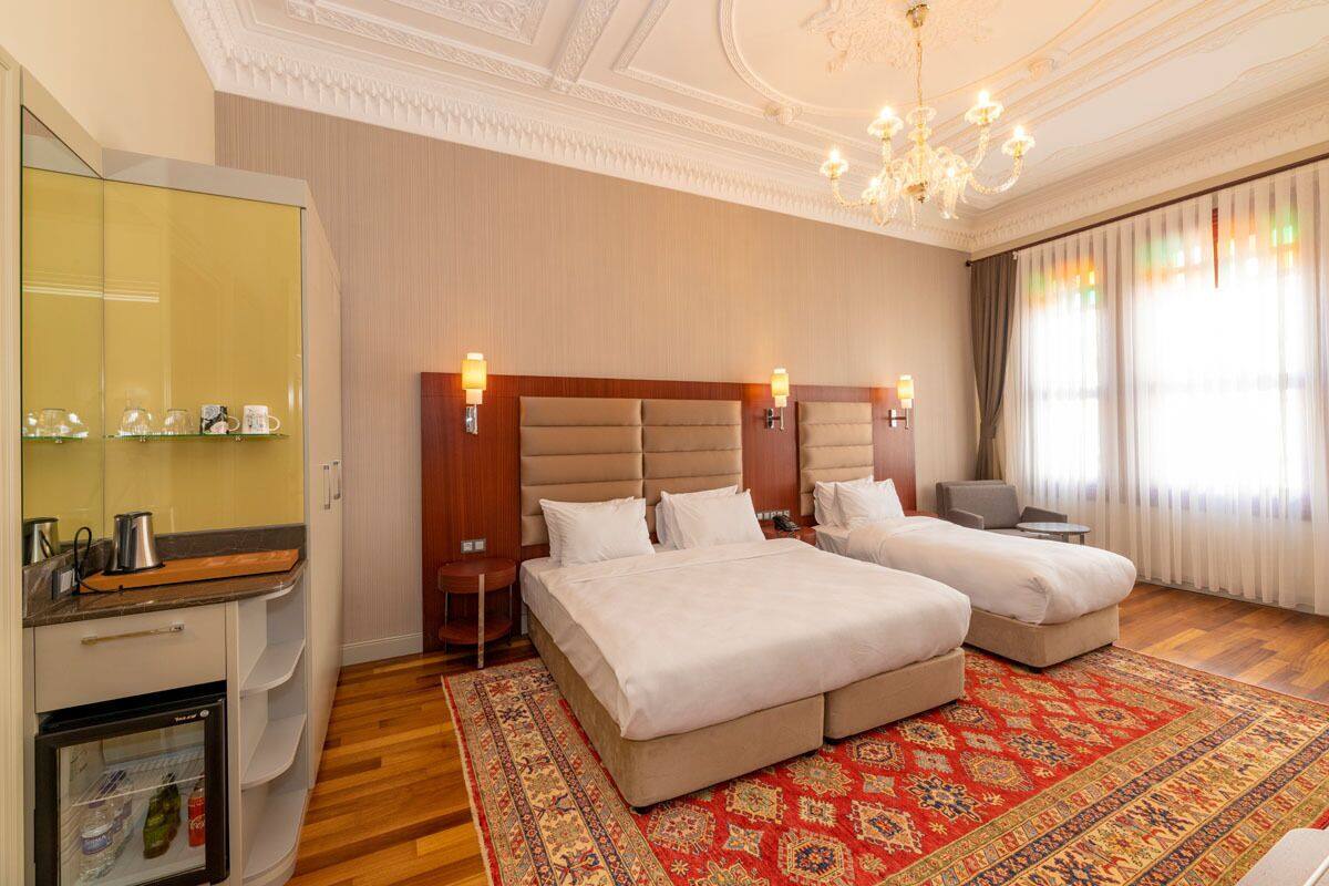 A hotel room with a large bed, a smaller bed, a minibar, a desk, and a large window with curtains.