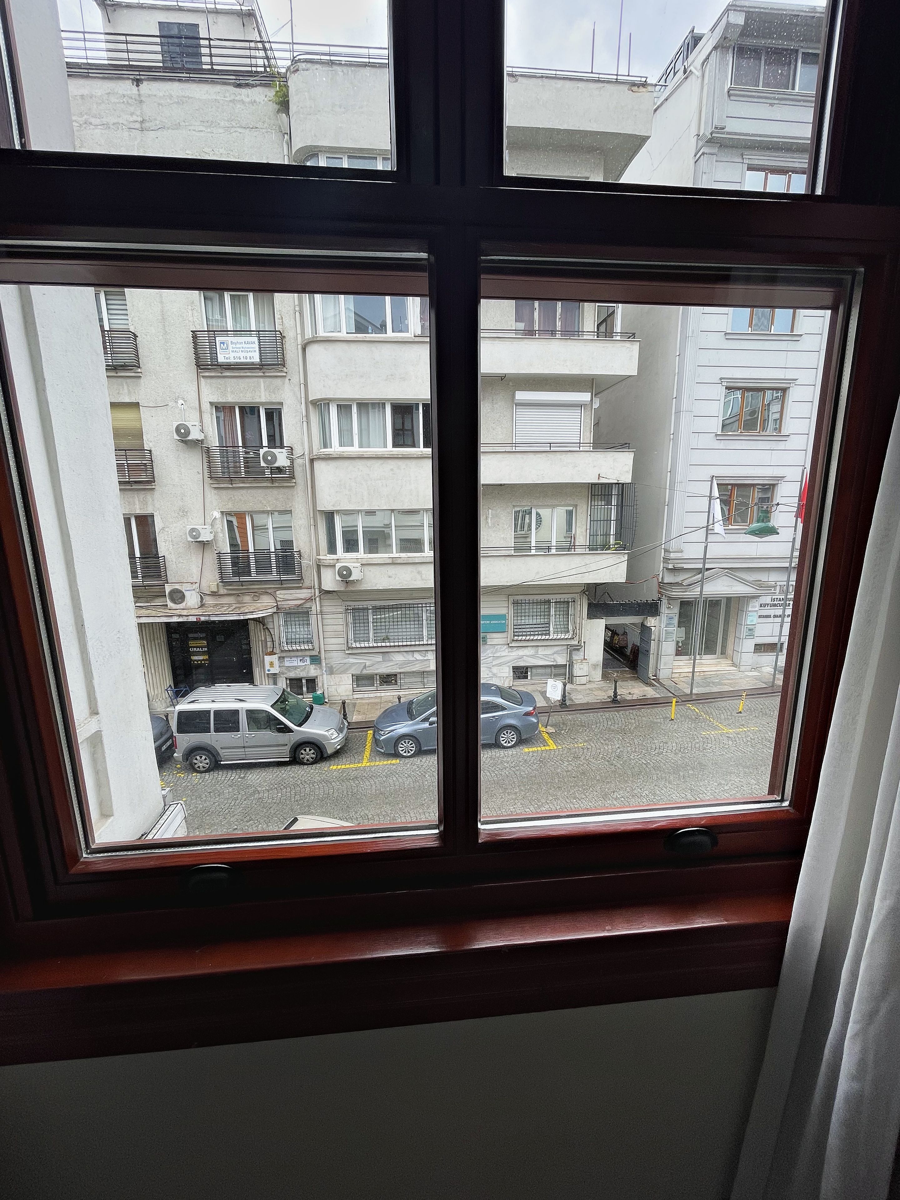 Blick durch ein Fenster mit vier Fenstern auf eine Straße mit parkenden Autos und mehrstöckigen Gebäuden.