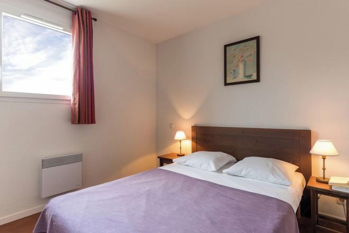 A hotel room with a bed, two bedside lamps, a window with curtains, a radiator, and a painting on the wall.