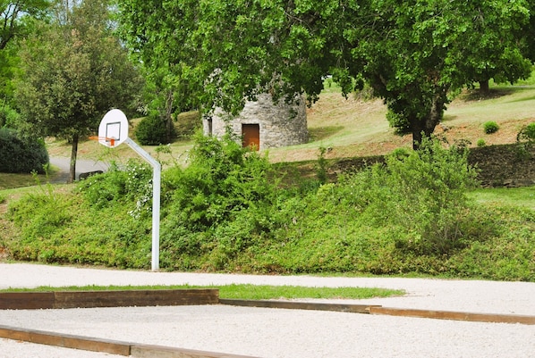 Uma quadra de basquete com um aro, cercada por vegetação e um edifício de pedra com uma porta.